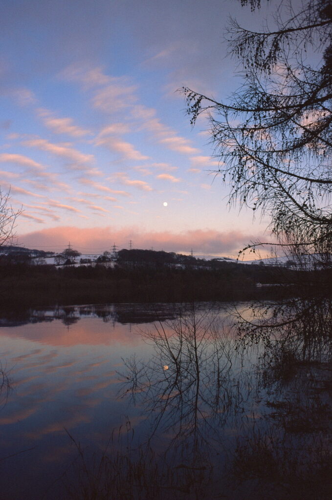 Pale blue sky with pink tinged clouds are reflected in the still water of a reservoir.